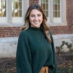 Headshot of brown-haired female staff member wearing a green sweater with a brick building in the background.