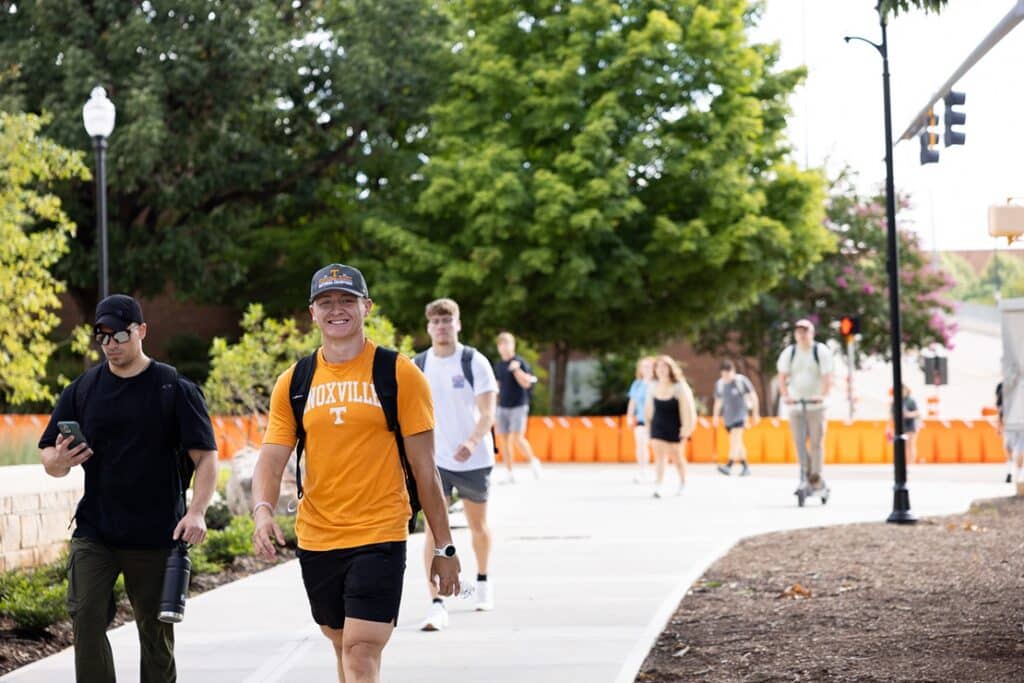 A UT student wearing an orange Knoxville shirt, smiling and walking on a sidewalk toward the camera with other students behind him.