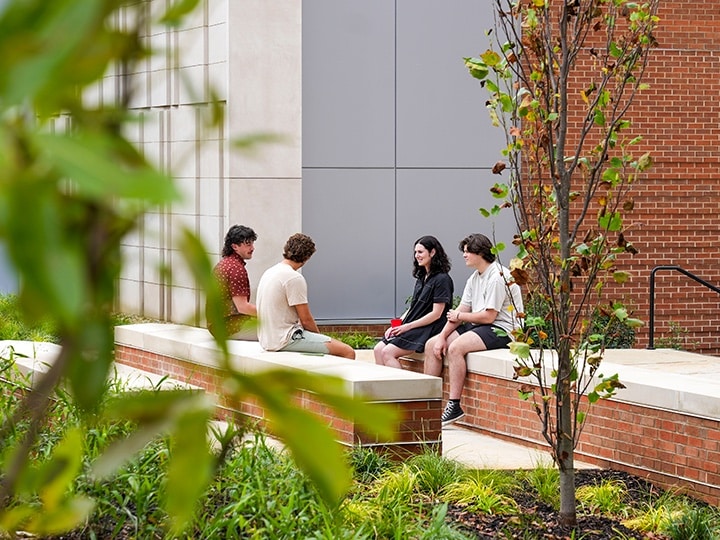 Four UT students sitting on stone structures outside the Agriculture and Natural Resources Building surrounded by plants and a tree.