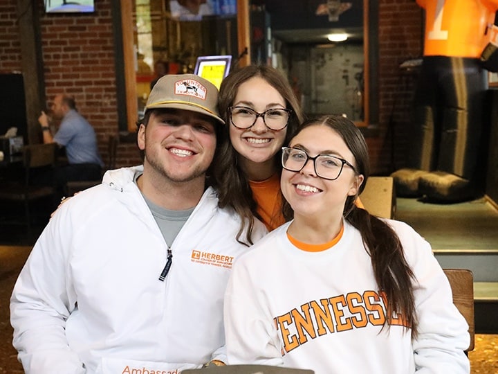 Three Herbert students wearing UT clothing and smiling in a restaurant.