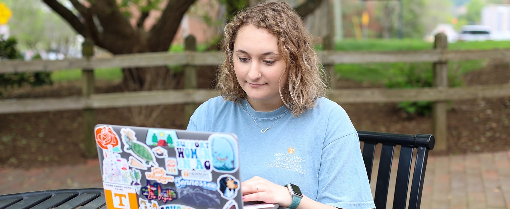 Female student sitting outside and wearing a blue shirt and "J" neckless on her computer that's covered in stickers.