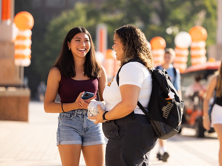 Two students smiling on a college campus sidewalk with orange and white balloon towers behind them.