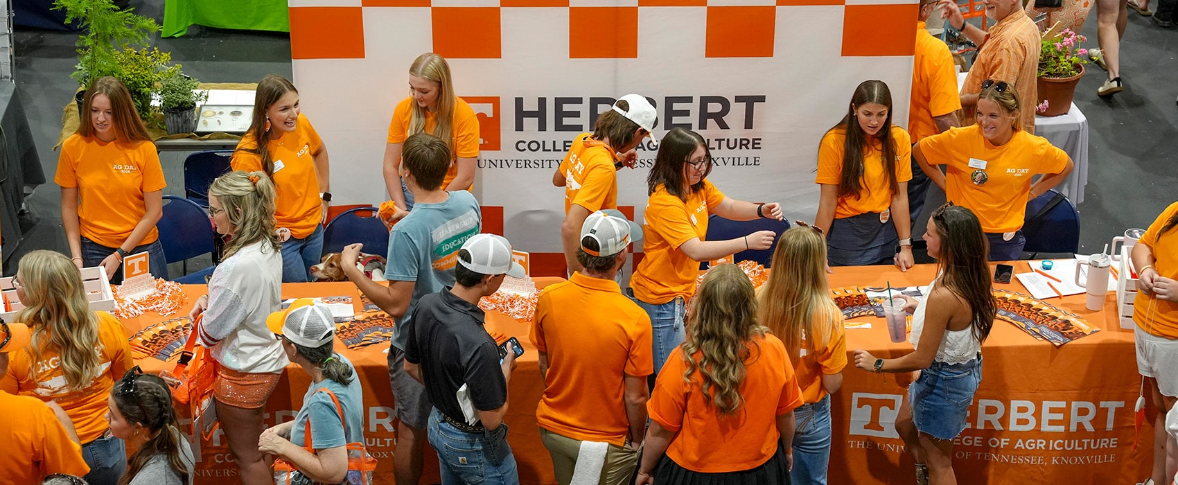 Herbert College of Agriculture students wearing matching orange shirts handing out swag at an event with a large checkerboard sign with the college logo on it behind them.