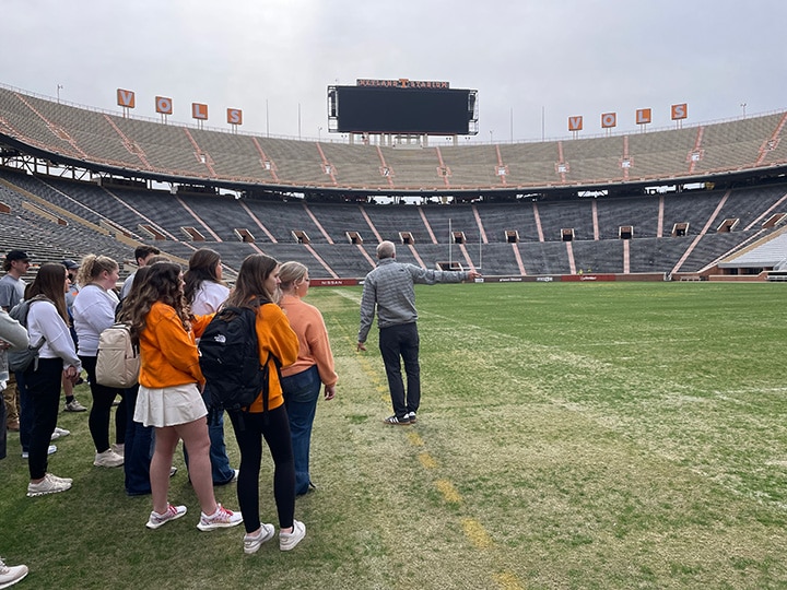 A professor with a group of students on the field at Neyland Stadium pointing to the turf grass.