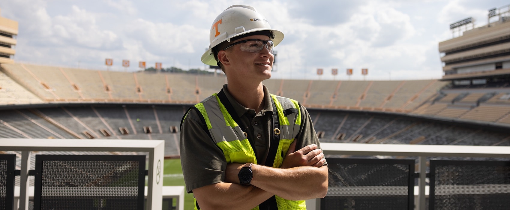 Construction science and management student standing with arms crossed in Neyland Stadium wearing a work vest, glasses, and a hard helmet with the Power T on it.