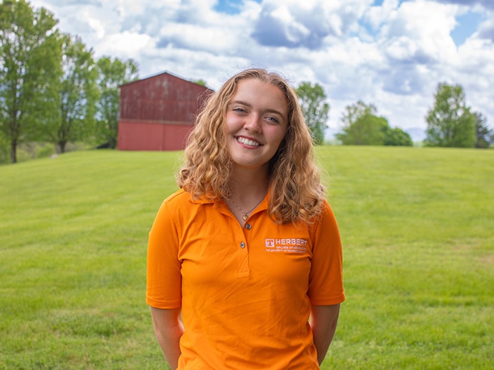 Headshot of Britta Gaetjens wearing a Herbert polo in a field with a red barn in the background.