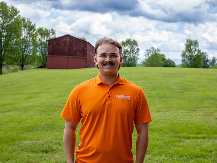 Headshot of Grey Johnson wearing a Herbert polo in a field with a red barn in the background.