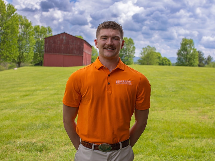 Headshot of Max Baldwin wearing a Herbert polo in a field with a red barn in the background.
