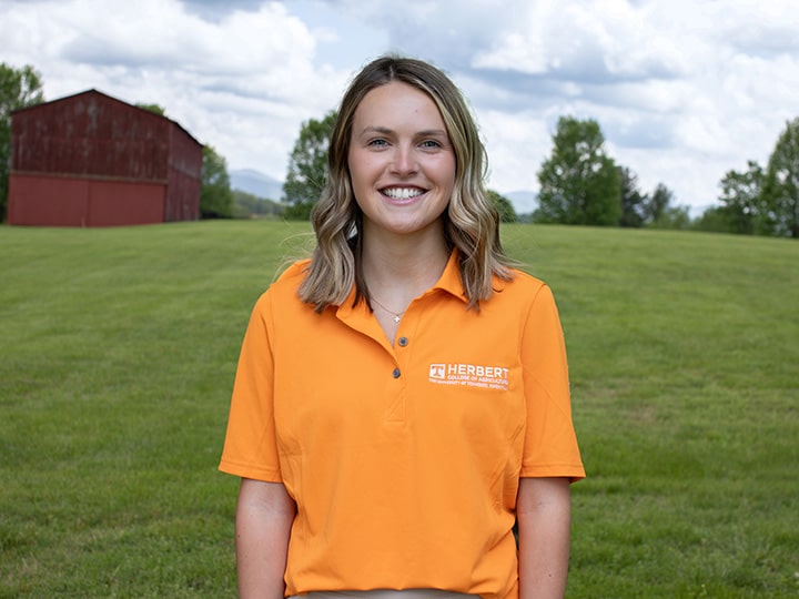 Headshot of Sarah Grace Kosek wearing a Herbert polo in a field with a red barn in the background.