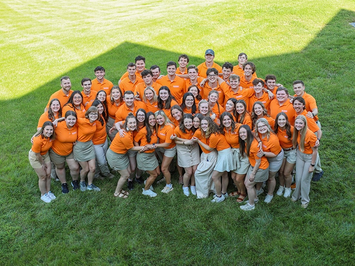 A group of students in matching orange polos hugging each other in a grassy field.