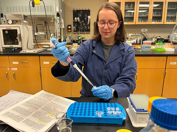 A student with glasses and wearing a blue lab coat using lab tools in a lab setting.