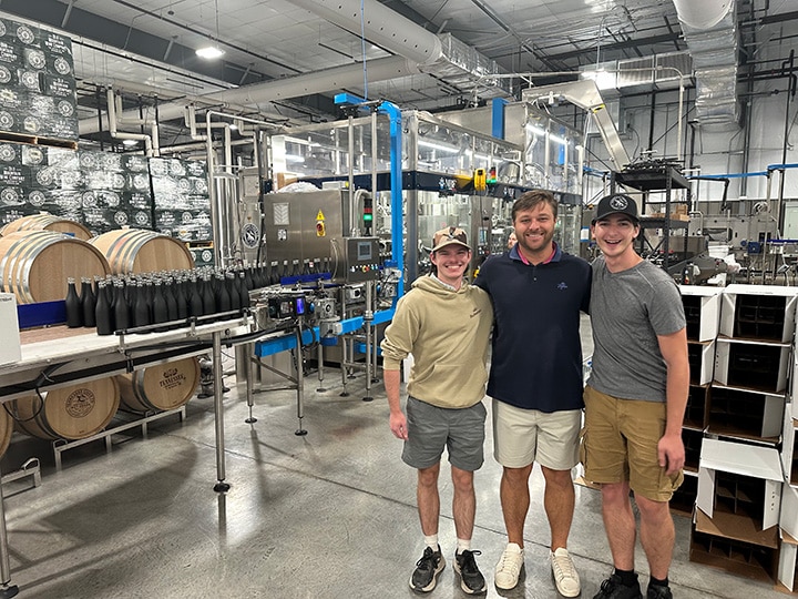 Three individuals in the back of a facility where wine is produced, surrounded by silver machinery, barrels, and bottles.