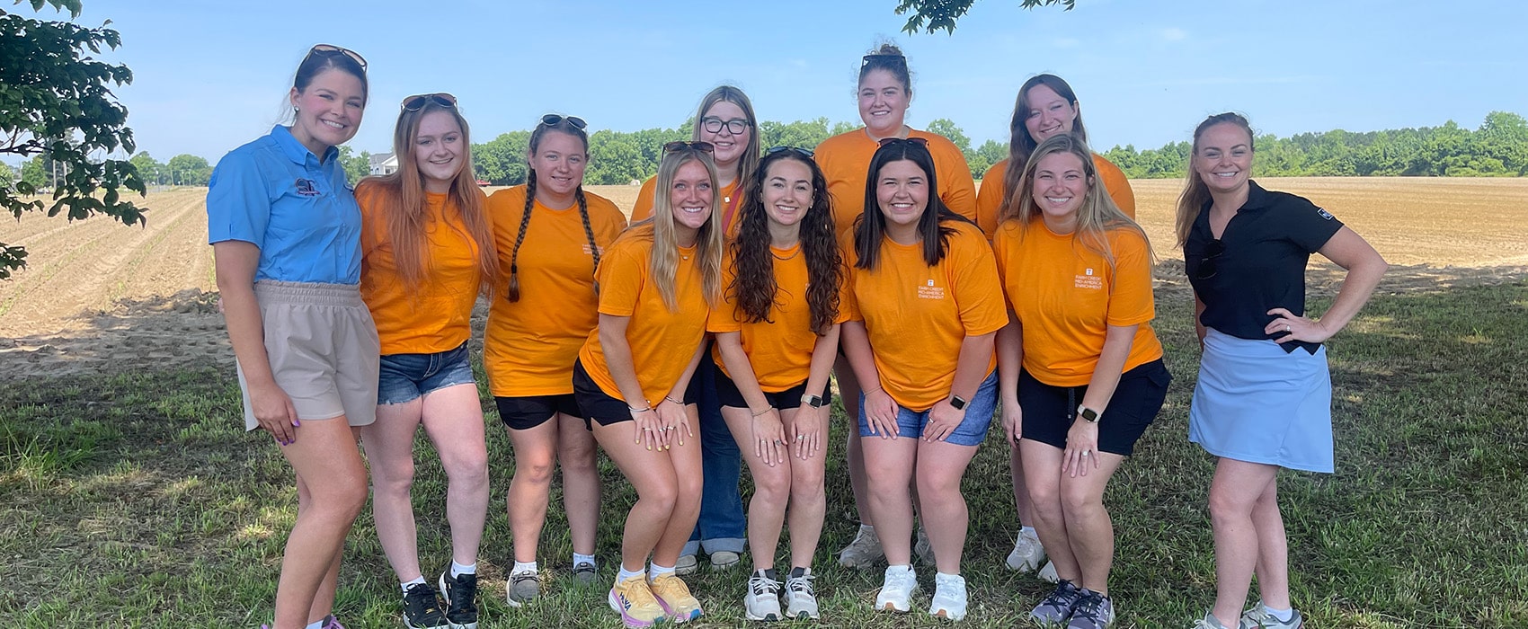 Group of college students and advisors posing for a photo in a field with most wearing orange shirts.