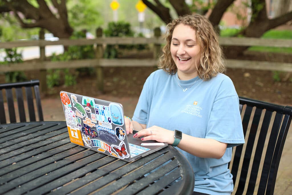 A student on her computer at a table outside, laughing and wearing an Agricultural Leadership, Education & Communications T-shirt.
