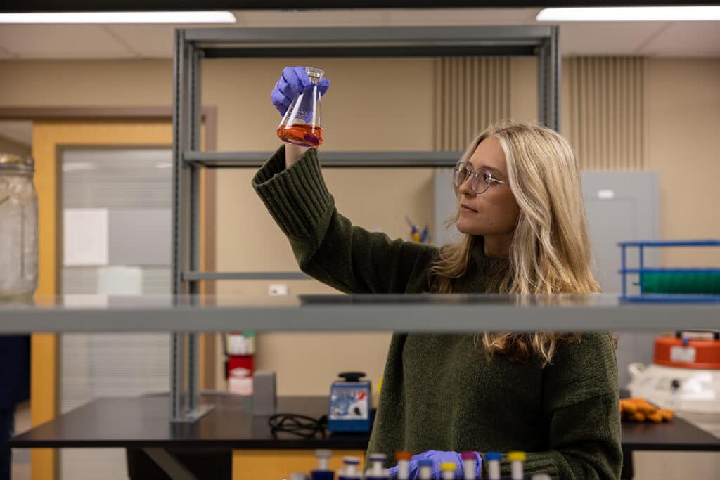 An animal science student wearing a green sweater and holding up an Erlenmeyer flask in a lab setting.