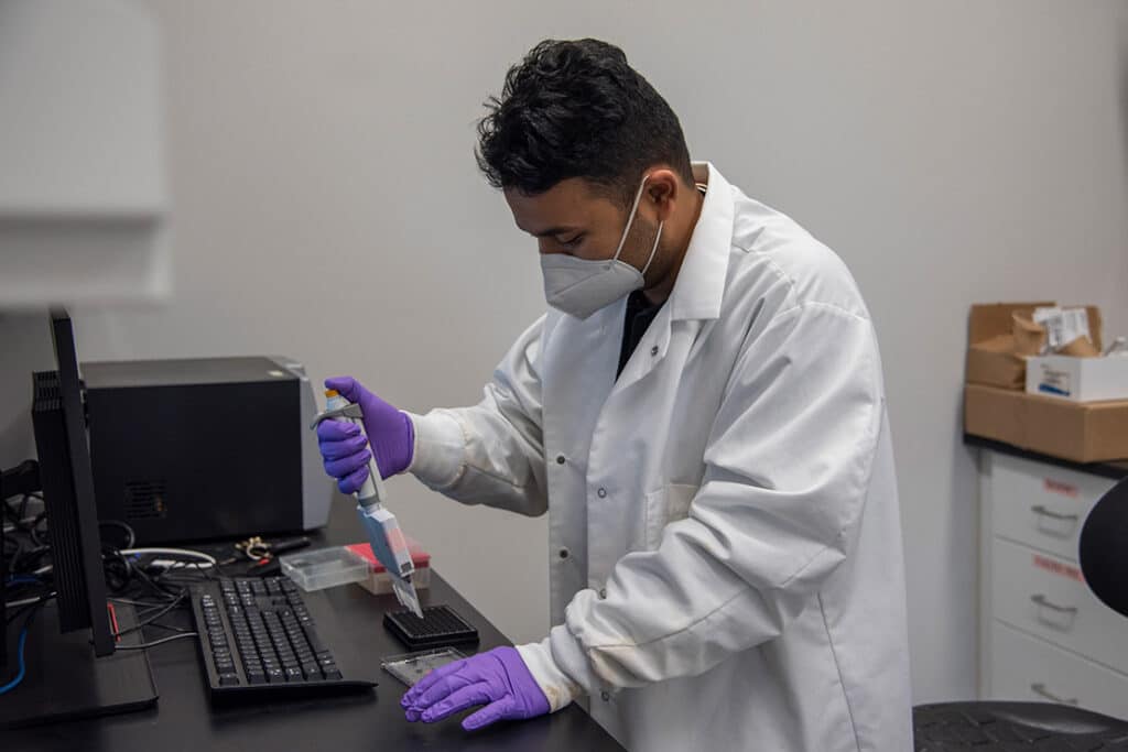 A graduate student wearing a white lab coat, purple gloves, and a face mask, conducting research near a computer.