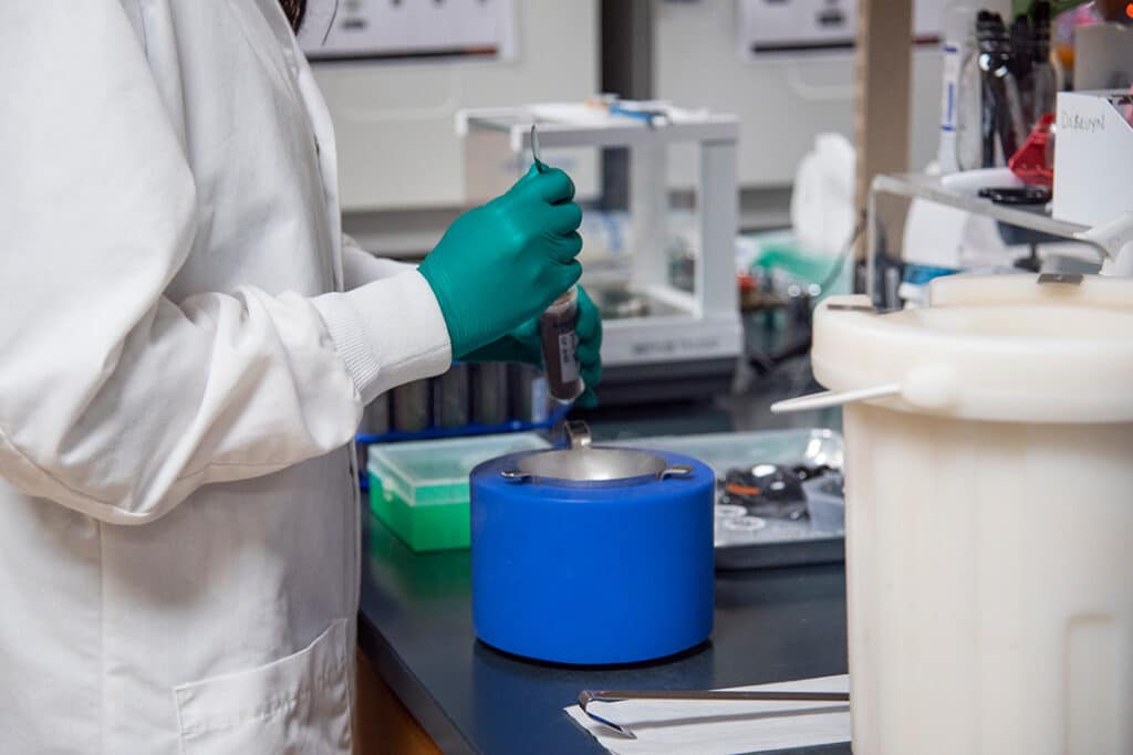 A graduate student wearing a white lab coat and green gloves conducting research using a test tube and a blue cylinder-like piece of equipment in a lab.