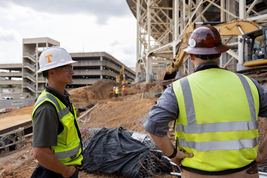 A construction science and management student wear a hard hat with a power T on it, talking to a construction professional outside Neyland Stadium.