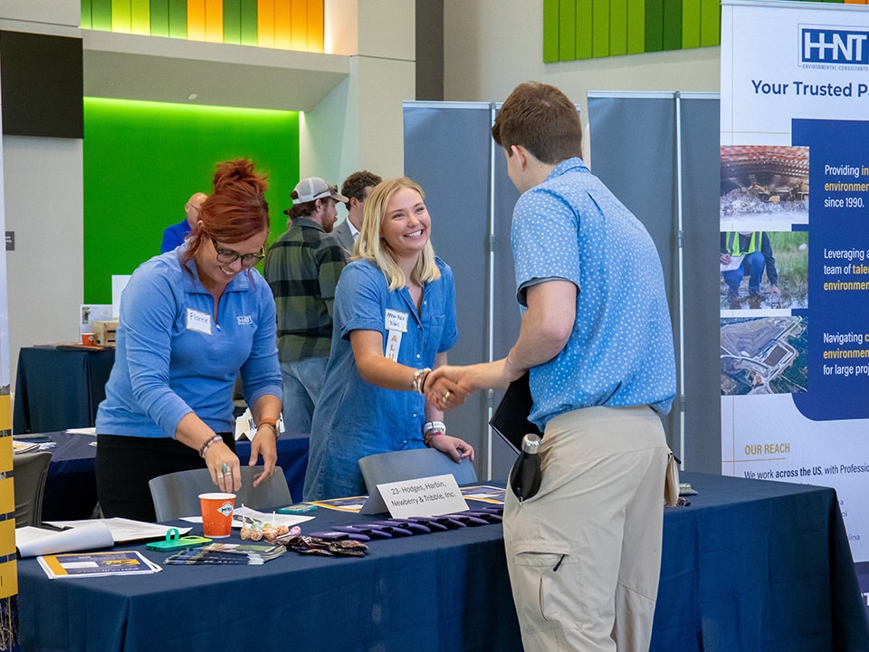 A student wearing a blue polo shirt shaking the hand of an employer at a career fair, also wearing blue and smiling.