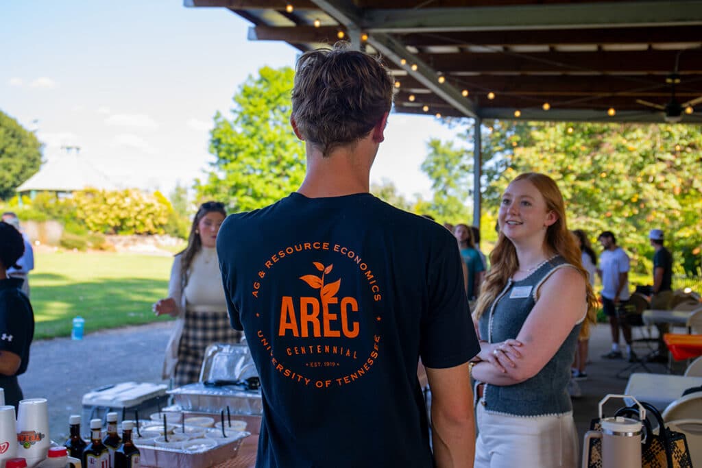 The back of a student wearing a black "AREC" shirt, talking to a professor at an outdoor picnic.
