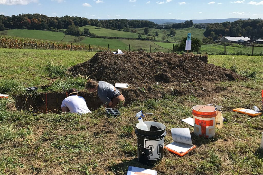 Students working in a hole next to a large pile of soil with black and orange power T buckets nearby.