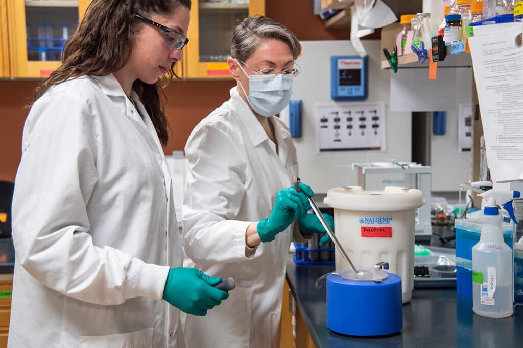A professor and a student wearing white lab coats and rubber gloves as they conduct research in a lab.