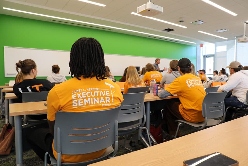 The backs of students in a classroom with a green front wall, wearing orange shirts that say "James L. Herbert Executive Seminar Series."