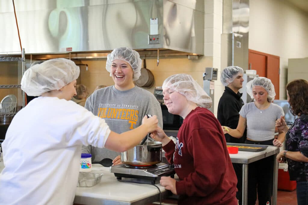 Students in a food science lab wearing hair nets and stirring something in a pot over a burner.