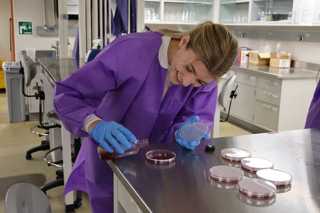 A student wearing a purple lab coat and blue rubber gloves, smiling, and pouring a liquid substance into a petri dish.