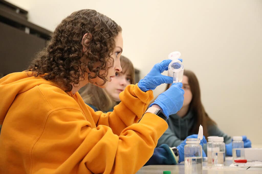 A student wearing an orange hoodie pouring a clear liquid into a test tube during a food science class.
