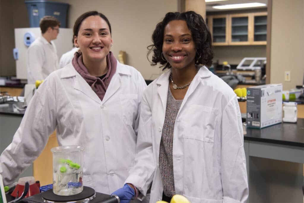 Two students in a food science lab wearing white lab coats, standing next to a beaker filled with test tubes.