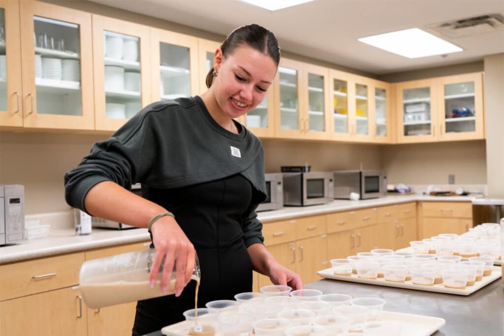 A student in the food science sensory lab pouring a brown substance into several sampling cups on trays.