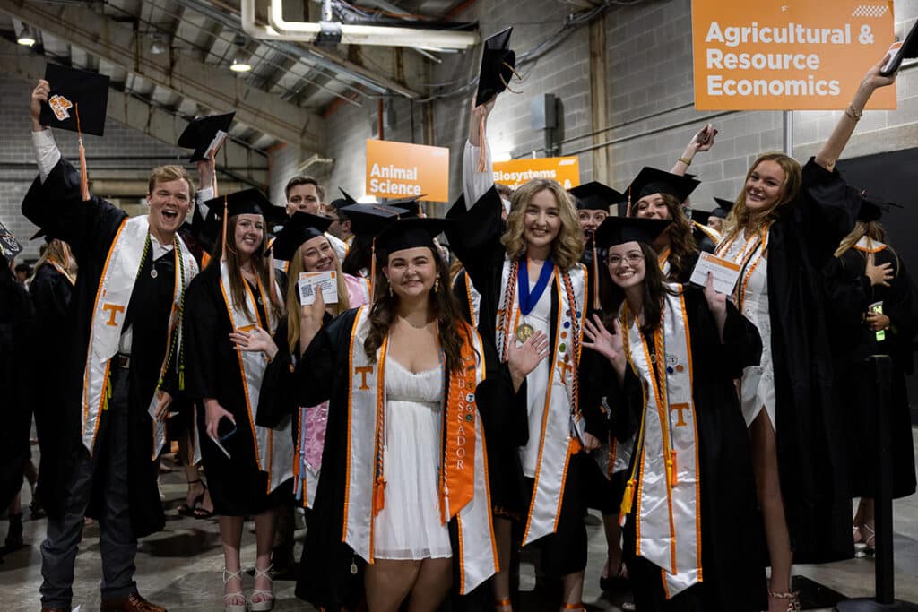 Students wearing graduation regalia standing next to an Agricultural and Resource Economics sign backstage at a commencement ceremony.