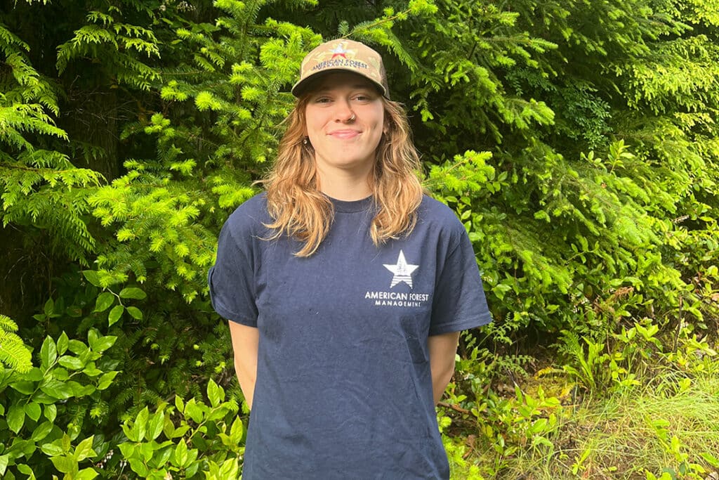 Finley Oldham wearing an American Forest Management shirt and standing outside in front of pine trees.