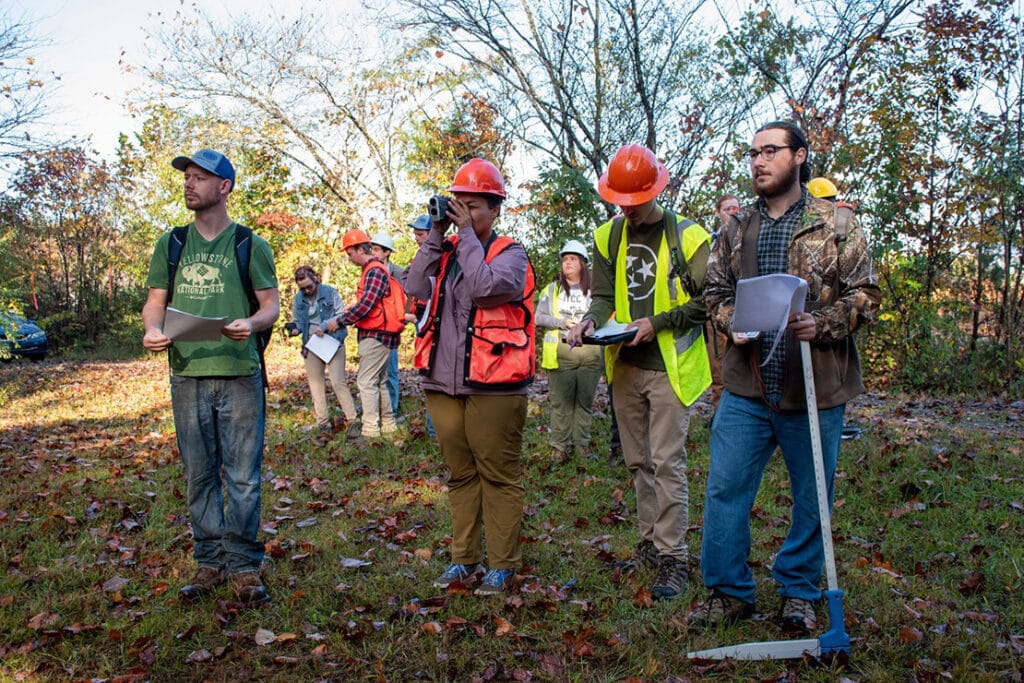 A group of forestry graduate students wearing bright hard hats and vests looking at trees in a wooded area.