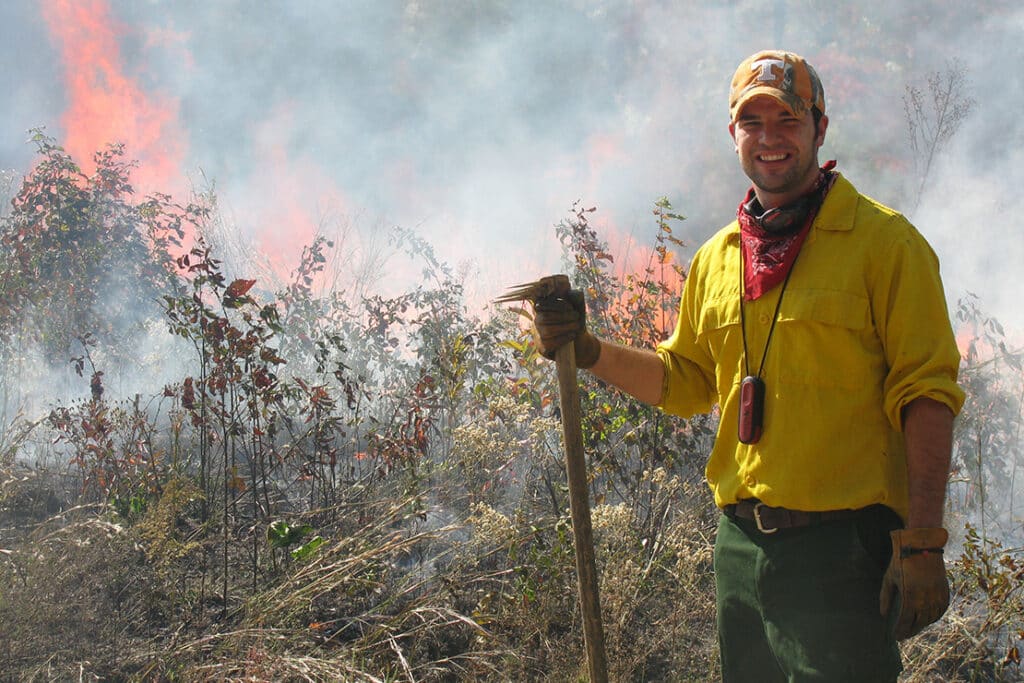 A natural resources student wearing bright yellow and an orange power T hat, smiling and working on controlled burns.
