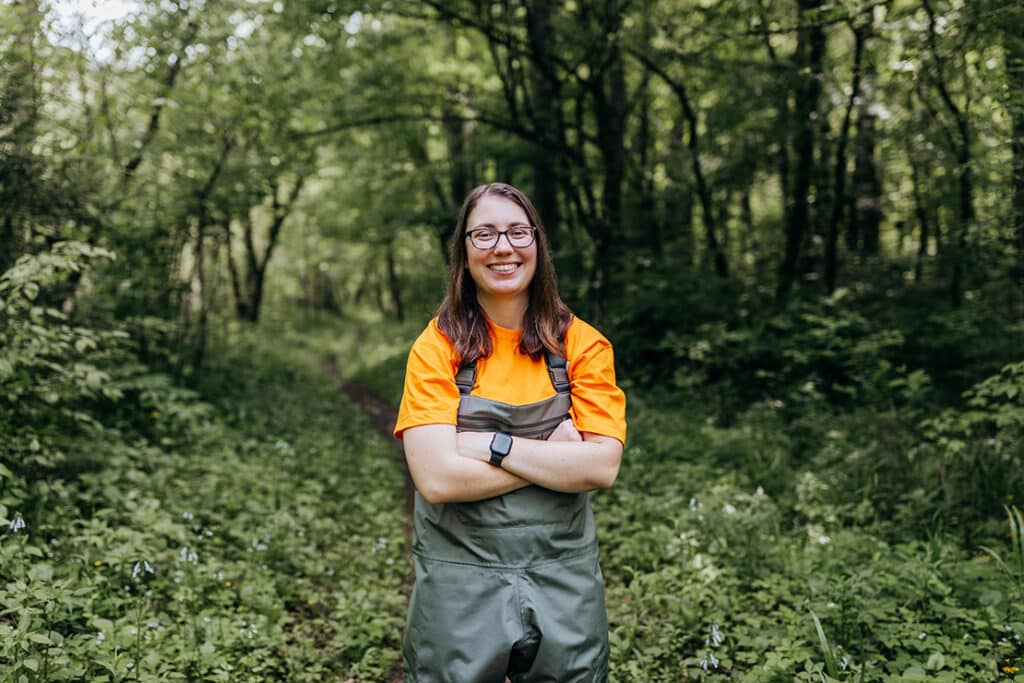 Wildlife and fisheries science student smiling and wearing waders and an orange shirt underneath in a wooded area.