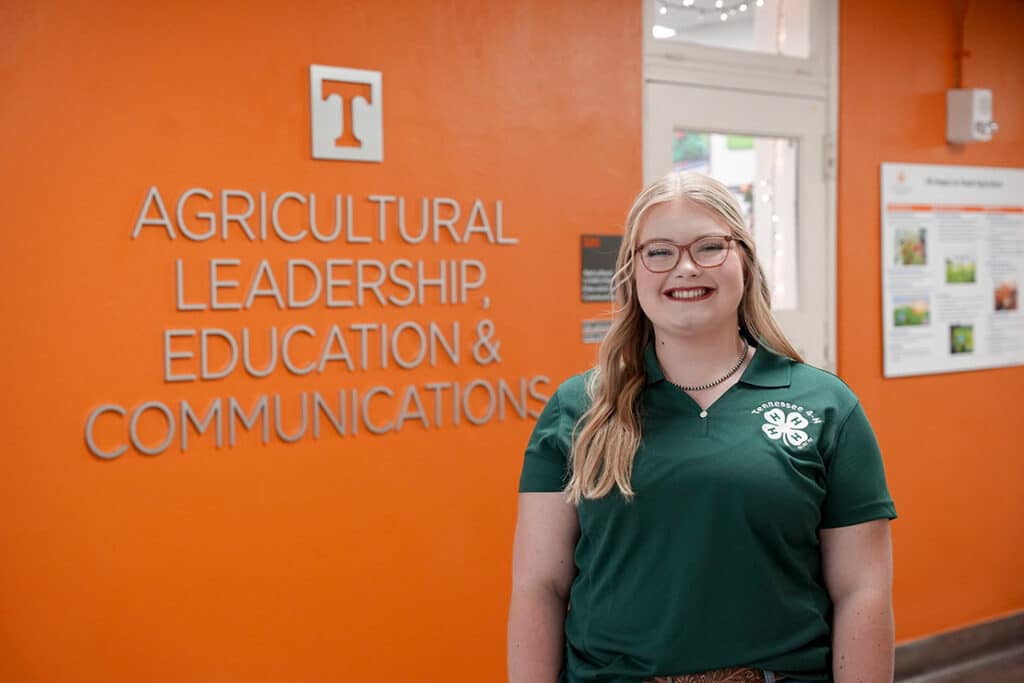 Student wearing a green Tennessee 4-H Day polo shirt, smiling and standing in front of an orange wall that says "Agricultural Leadership, Education & Communications."