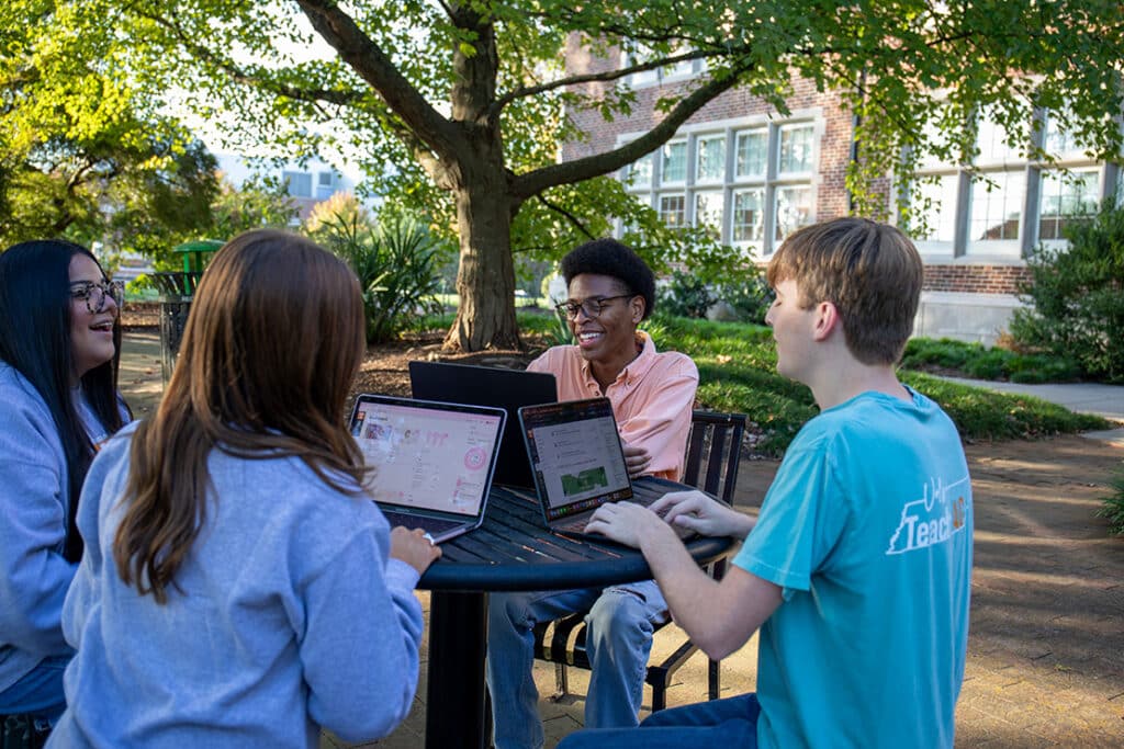 A group of students sitting at a table outside a brick academic building, smiling, and working on their computers.