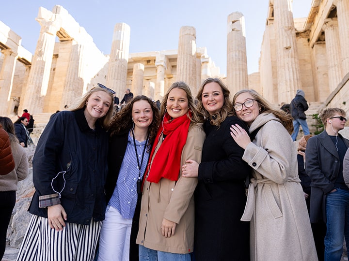 Five Herbert students smiling in front of the Parthenon on the Acropolis in Athens. They are dressed in warm, casual clothing, and the ancient structure with its tall columns is visible in the background.
