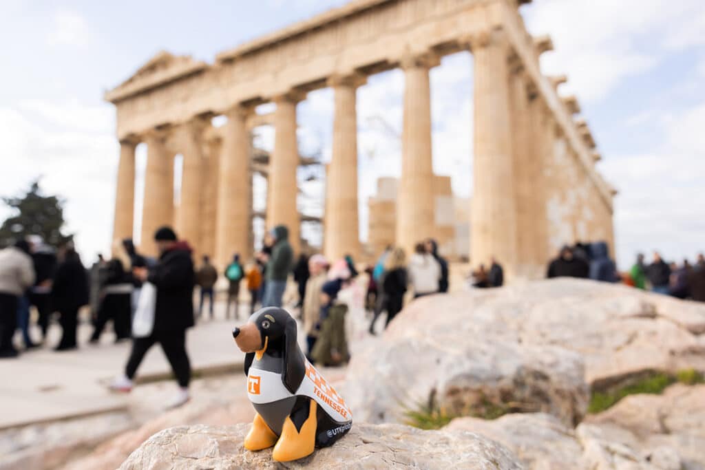 A figurine of Smokey, the University of Tennessee's mascot, is placed on a rock with the Parthenon in the background. People are visible walking around the monument.