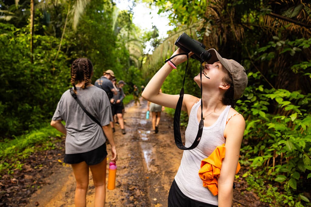 Student looking up through binoculars on a muddy trail in a jungle with other students walk ahead of them.