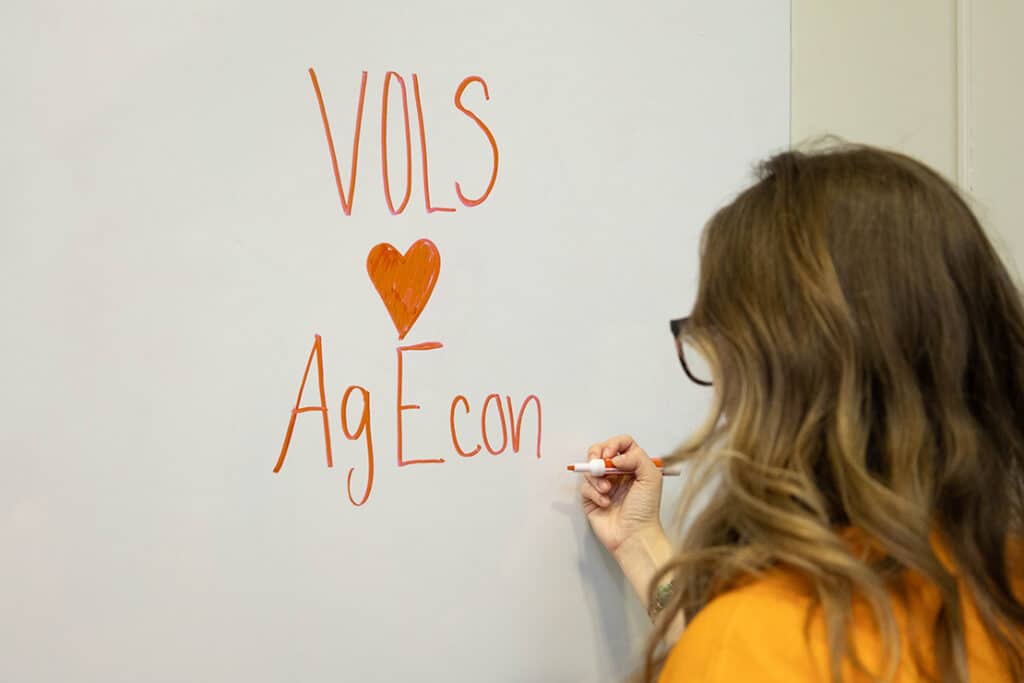 A student wearing an orange shirt writing on a white board with an orange marker, "VOLS (heart sign) AgEcon."