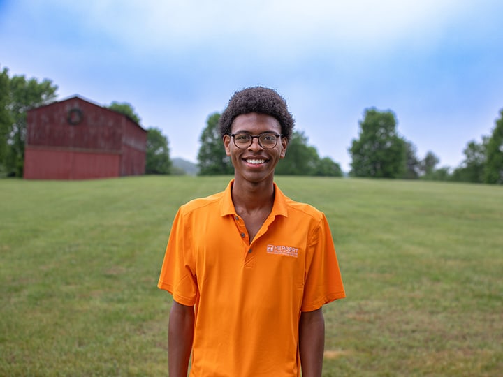 Headshot of Kendall Beard wearing a Herbert polo in a field with a red barn in the background.