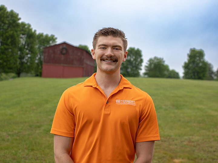 Headshot of Max Baldwin wearing a Herbert polo in a field with a red barn in the background.