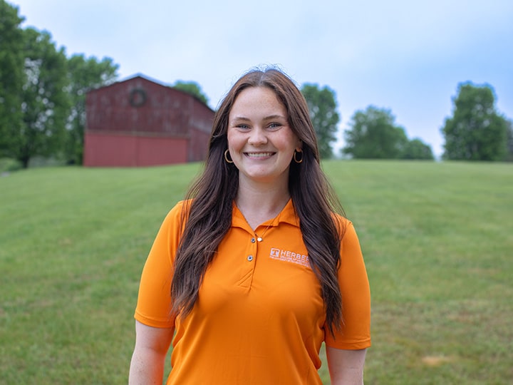 Headshot of Nora Mooney wearing a Herbert polo in a field with a red barn in the background.