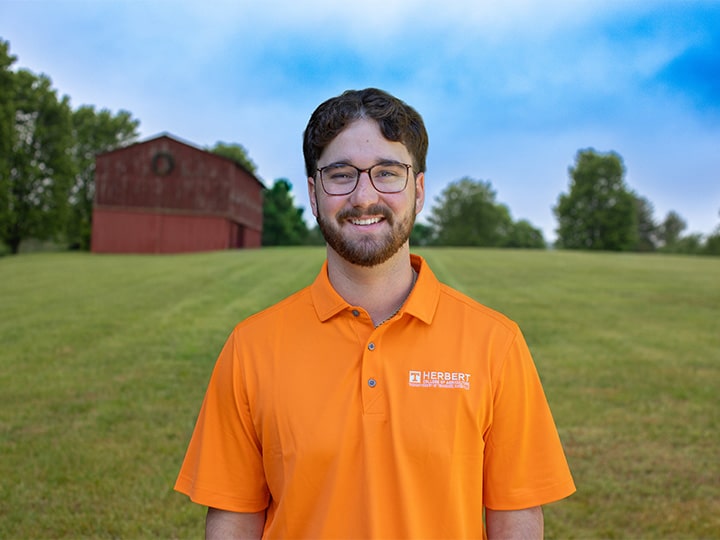 Headshot of Thatcher Scholar wearing a Herbert polo in a field with a red barn in the background.