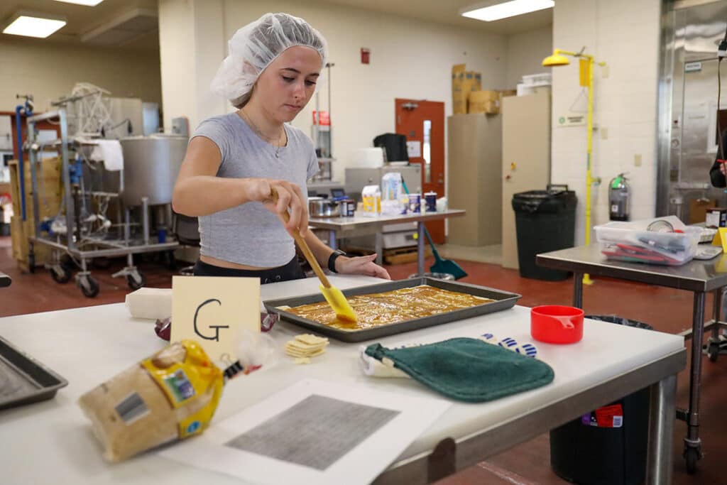 Student wearing a hair net, coating a sheet pan with a brown substance in a food science lab setting. Photo by Celina Menard.