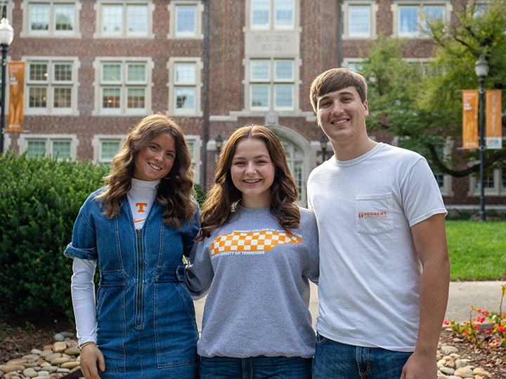 Three agricultural leadership, education, and communications students standing and smiling with their arms around each other outside the front of Morgan Hall while wearing UT Knoxville and Herbert attire. Photo by Celina Menard.