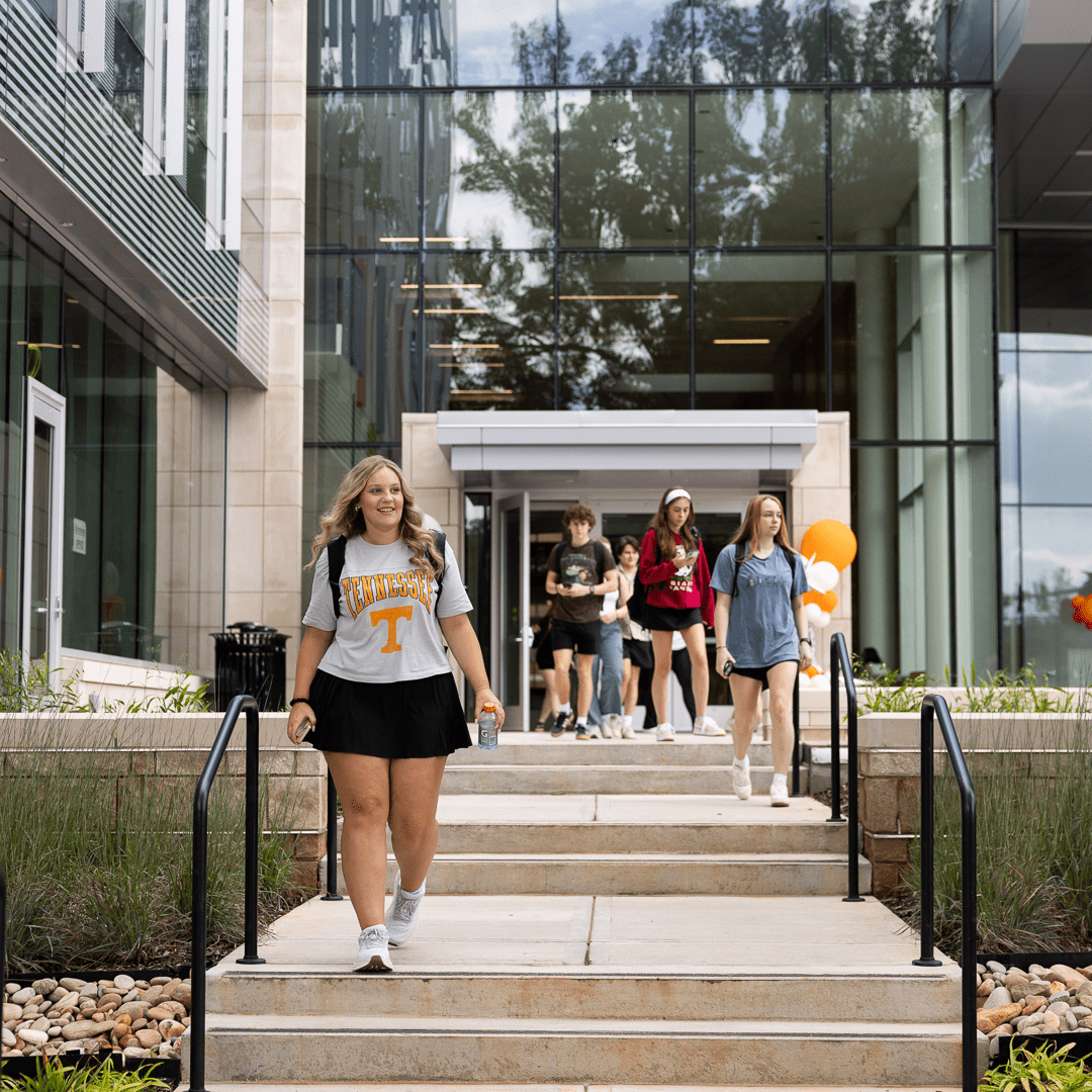 Herbert student wearing a black skirt and Tennessee t-shirt walking outside of a large glass academic building.